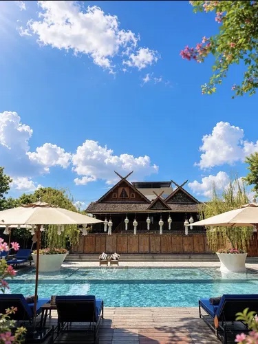 A teak Lanna pavilion with carved gable finials stands behind a long swimming pool, cream parasols and loungers along the near edge, bougainvillea and blue sky above.