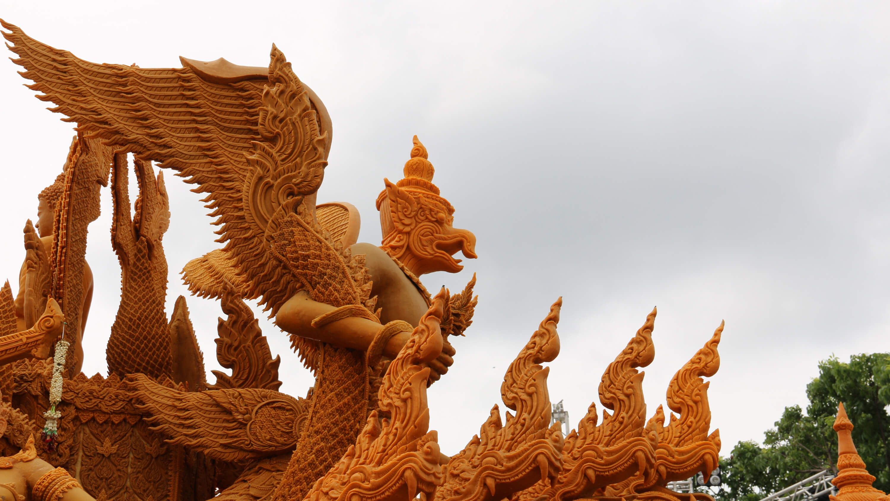 An ornate orange wax sculpture of a winged deity in traditional Thai carved style, flame-shaped finials along a roofline below an overcast sky.