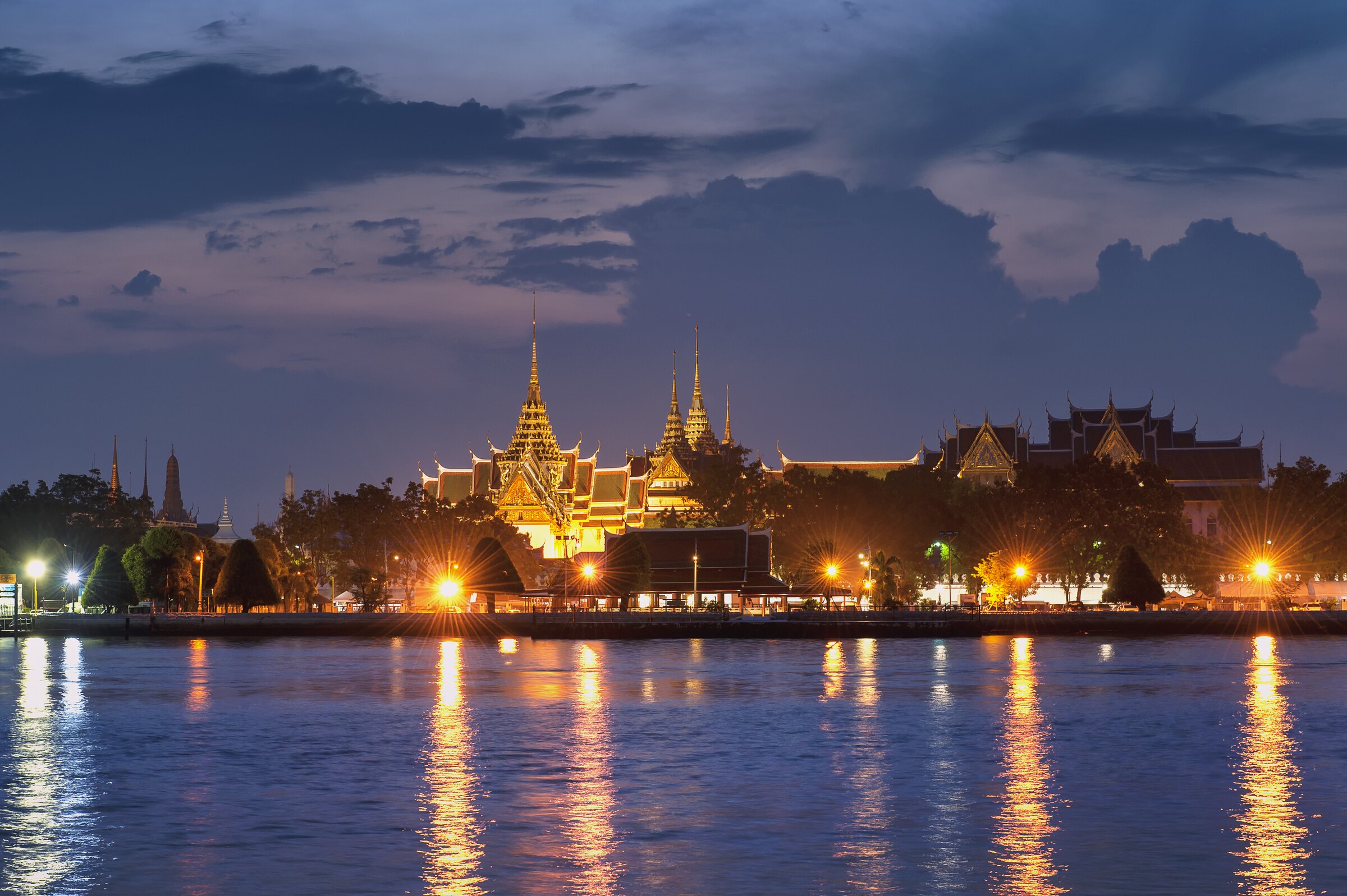 The Grand Palace lit at dusk across the Chao Phraya, its spires reflected in the river