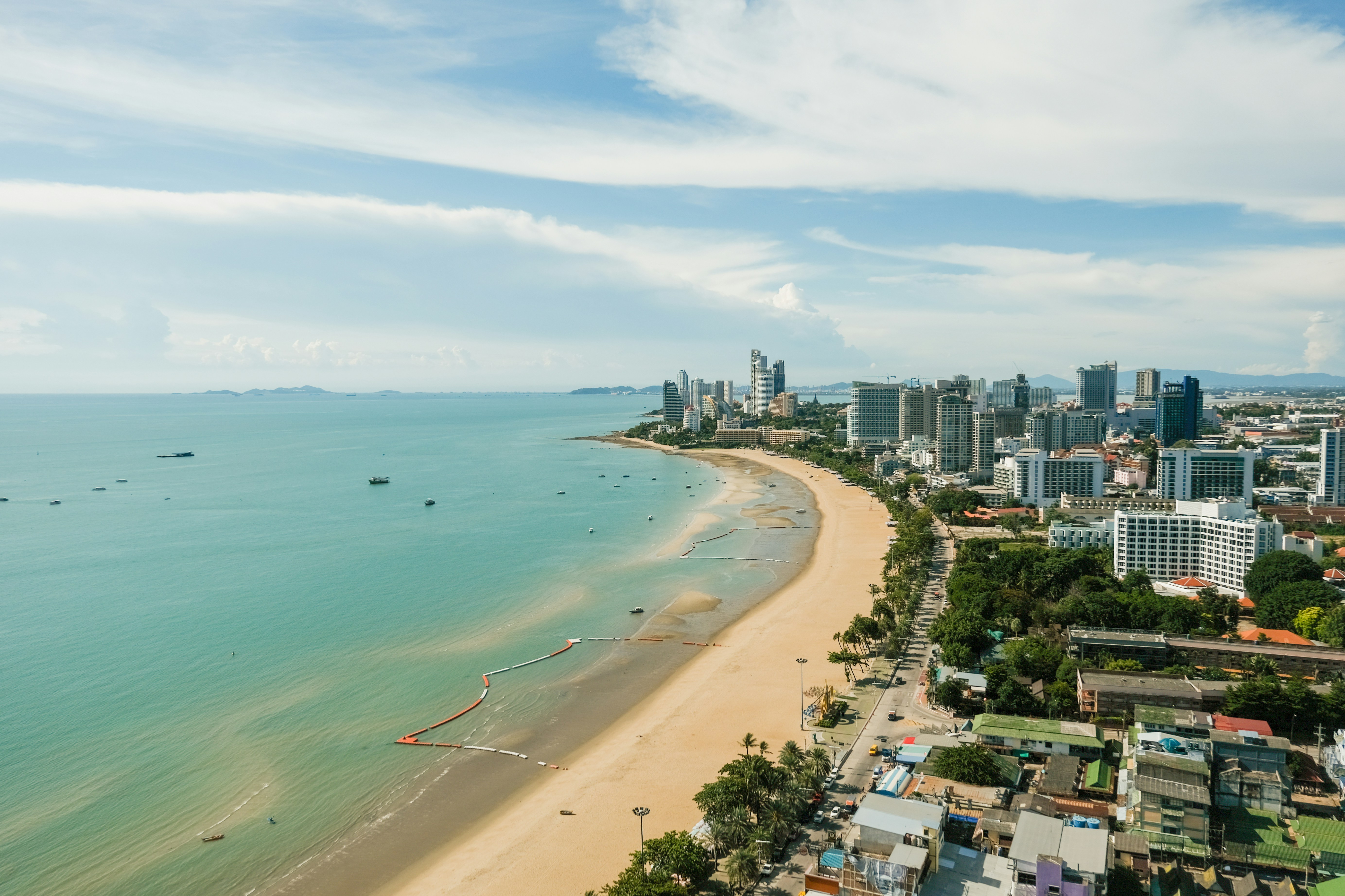 An aerial view along Pattaya's curving beach at midday, the high-rise skyline rising behind the shoreline and boats anchored offshore.