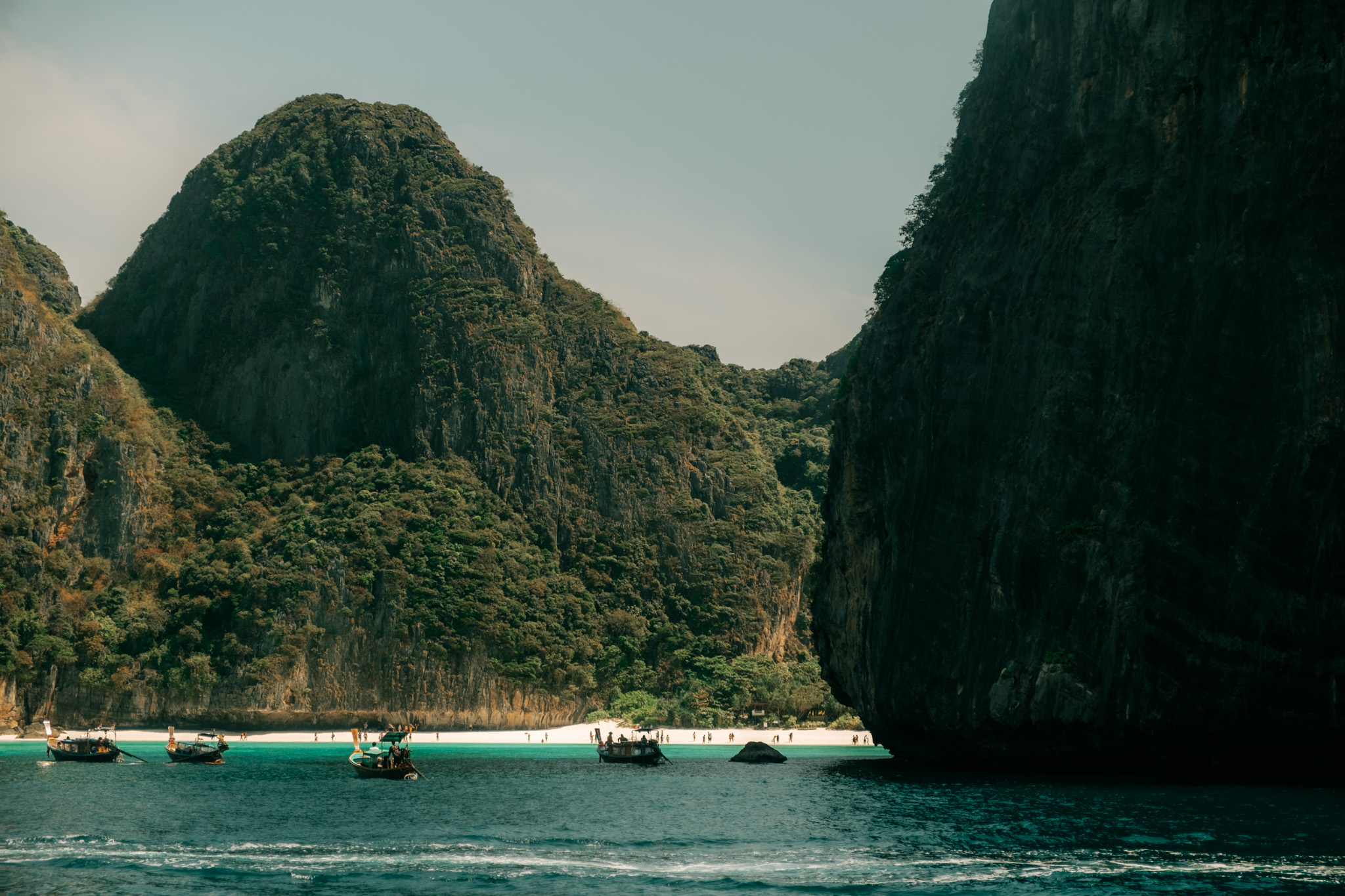 Steep forested limestone cliffs framing a sheltered cove, longtail boats moored in shallow turquoise water beside a white-sand beach.