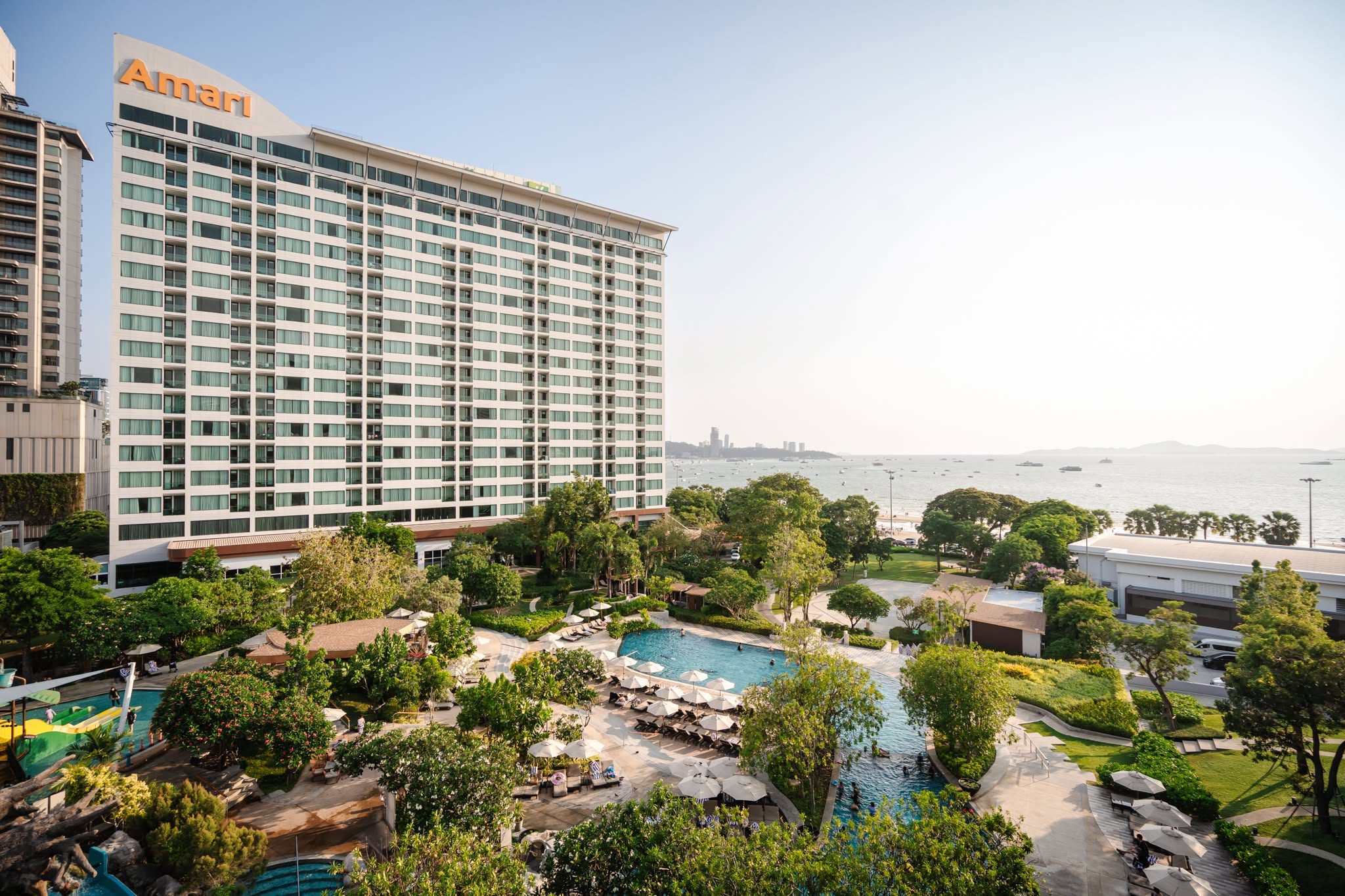 The hotel tower seen from its gardens, orange Amari signage at the roofline, a landscaped pool and water slide in the foreground with the sea beyond.