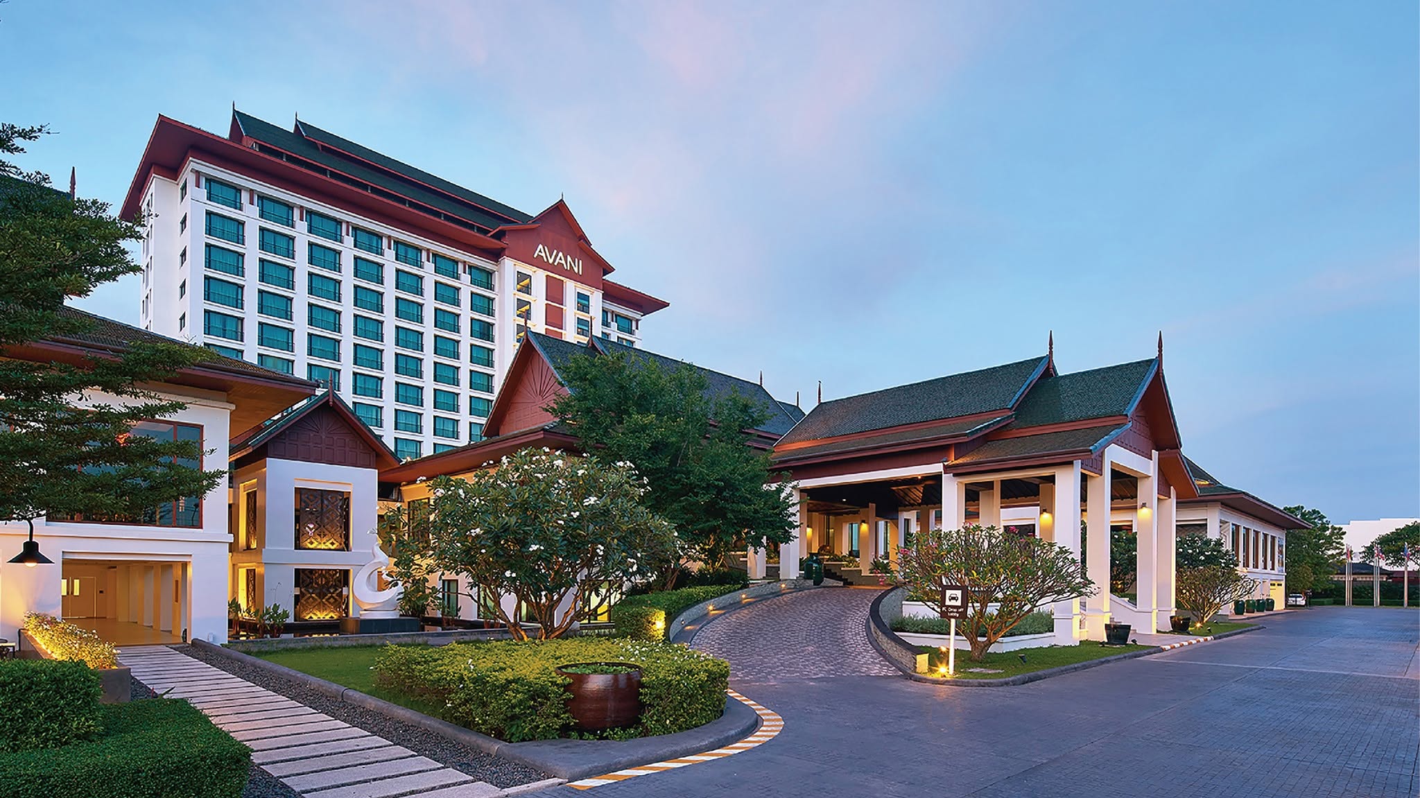 The hotel's tiered Lanna-roofed tower at dusk, AVANI signage lit at the crown, its porte-cochere and garden entrance in the foreground.