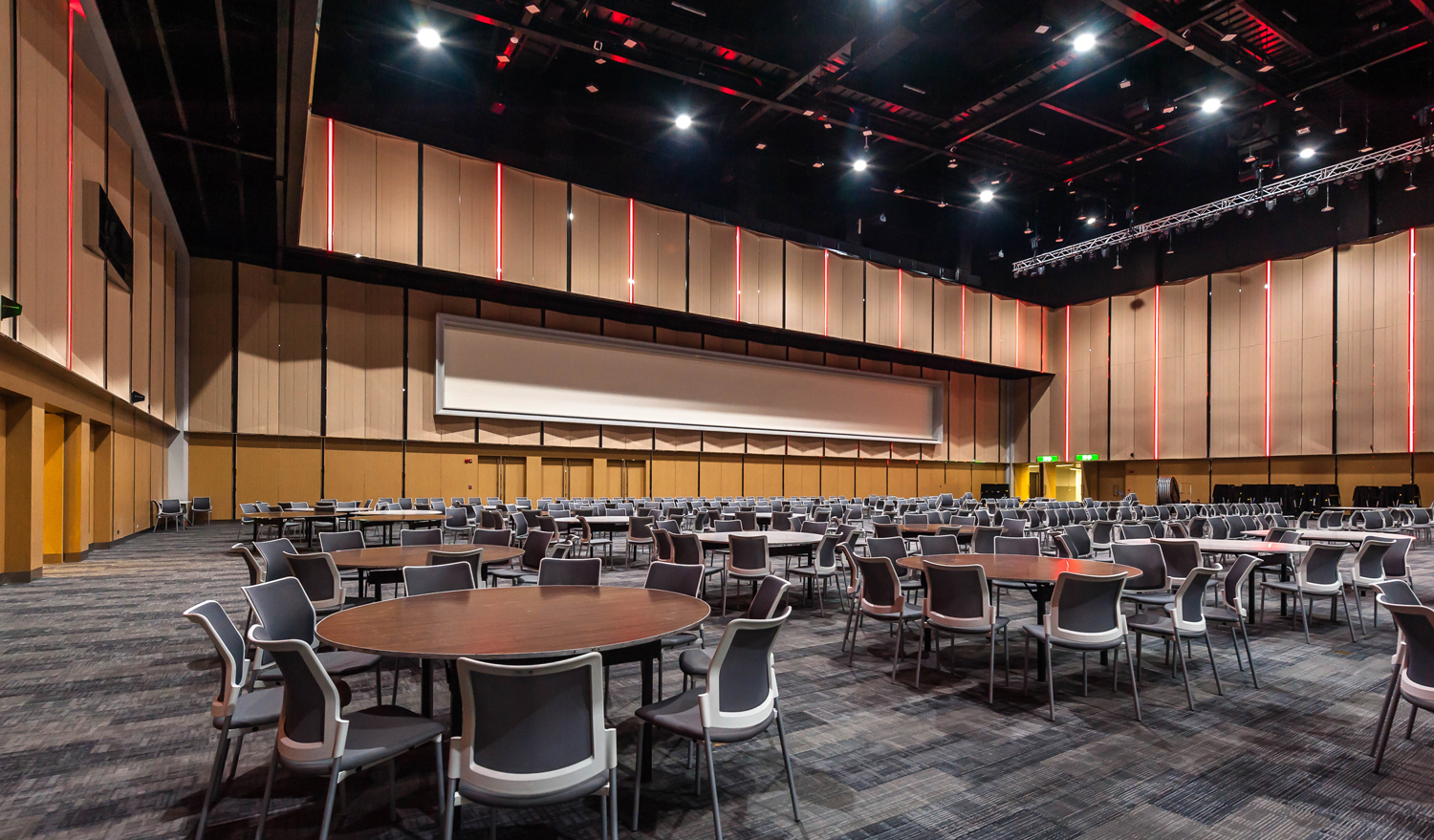 A function room set with round banquet tables and stacking chairs, timber-panelled walls lit by vertical red strips and a long projection screen at the far end.
