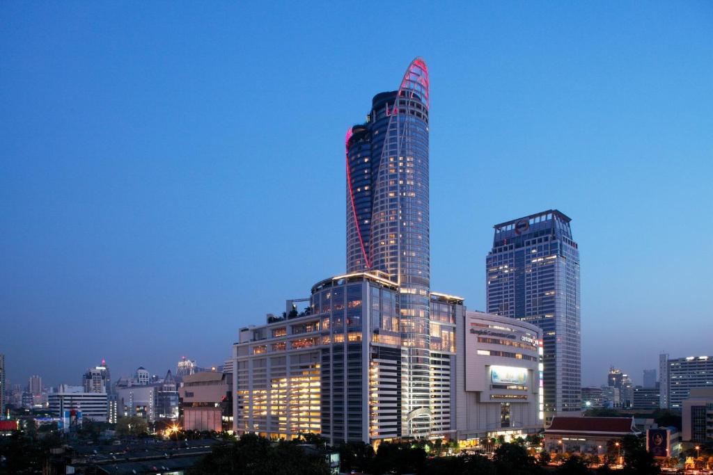 The hotel tower at dusk, a red illuminated arc tracing its curved crown above the CentralWorld complex, the lit city spread below.