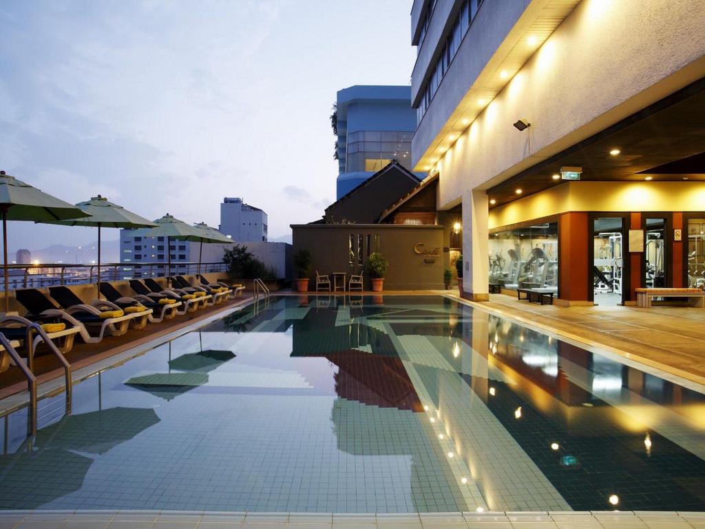 A rooftop swimming pool at dusk lined with loungers and umbrellas, the hotel's illuminated gym visible through glass to one side and the city skyline beyond.