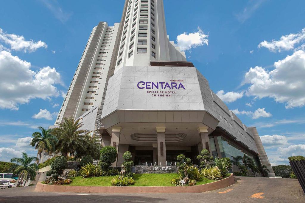 The hotel tower seen from its driveway, CENTARA RIVERSIDE HOTEL CHIANG MAI lettering above the porte-cochere and clipped topiary in the forecourt garden.