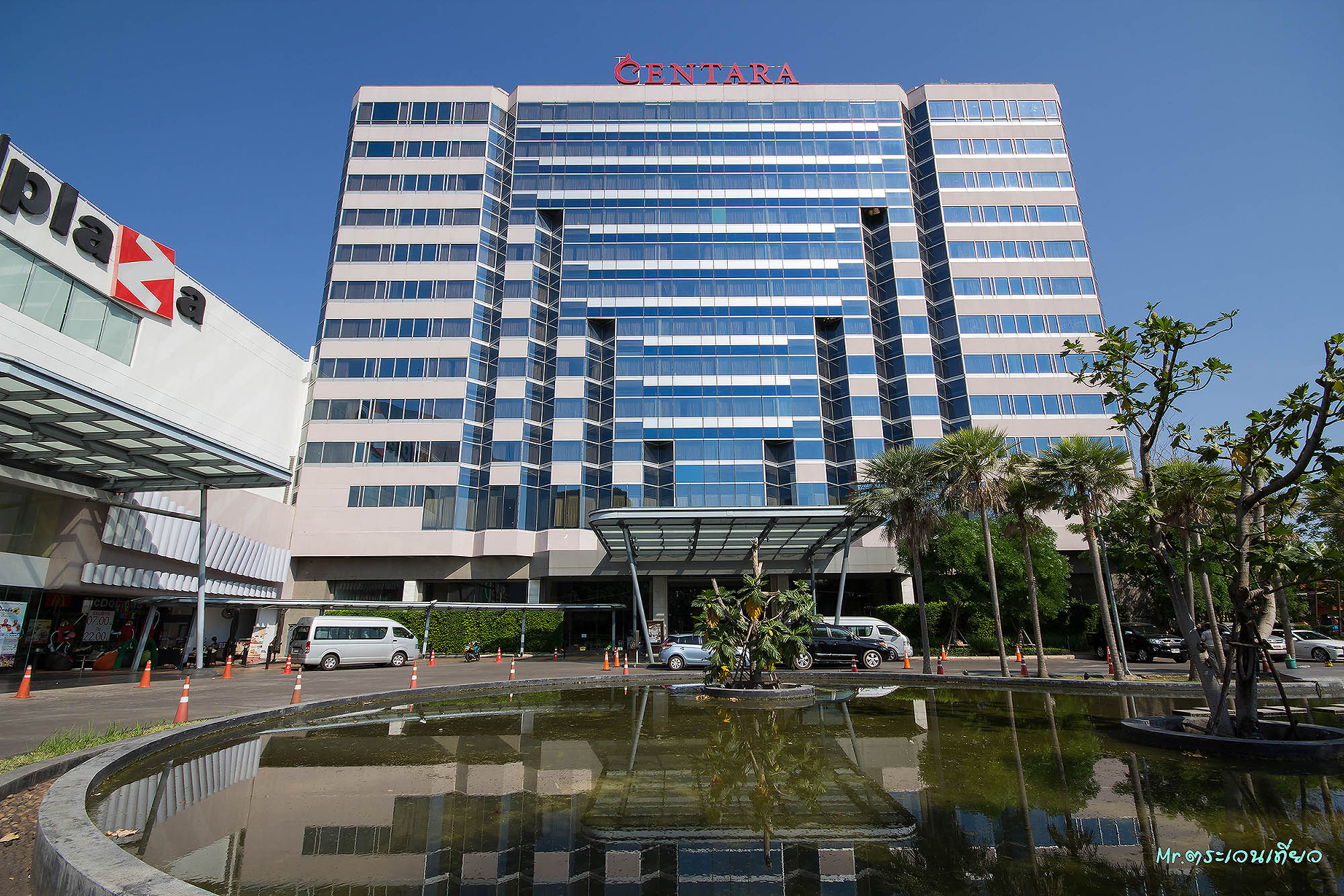 The hotel tower's glass and stone facade above the UD Town Plaza mall, red CENTARA signage at the crown and a landscaped fountain pond in the forecourt.