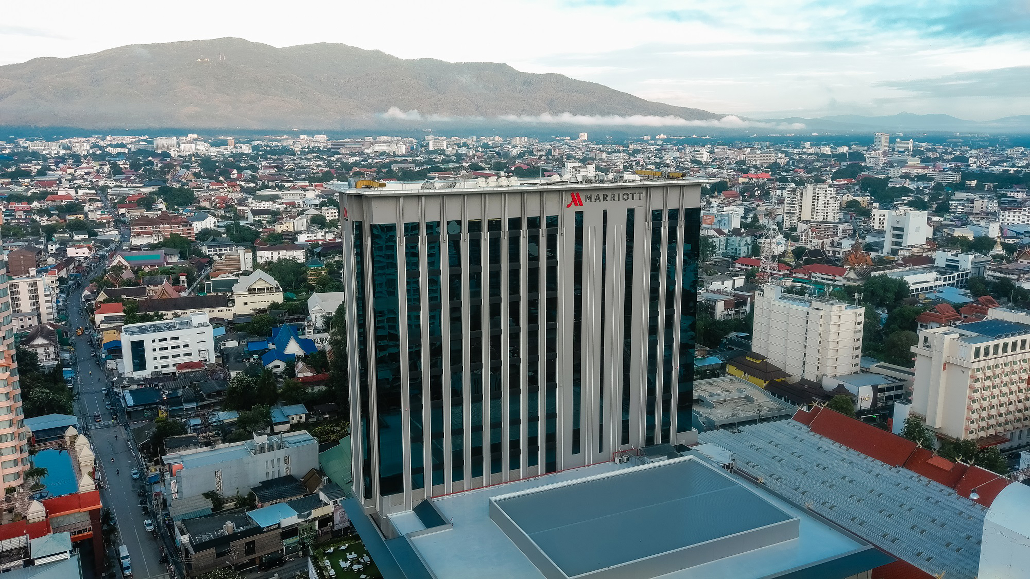 An aerial view of the tower with red MARRIOTT signage above its top floors, the old city and forested mountains spread behind it in early morning haze.