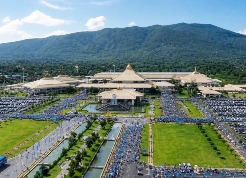An aerial view of the convention centre's tiered golden Lanna roofs against a forested mountain ridge, a large crowd gathered in blue across the forecourt and full car parks either side.