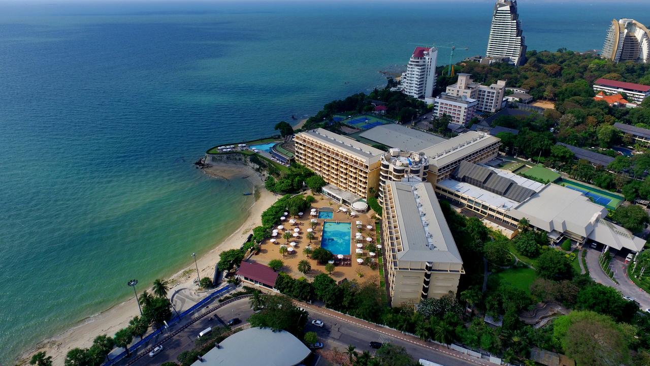An aerial view of the hotel's beachfront property on a small headland, pools and gardens beside the sand, tennis courts behind, and Pattaya's skyline further along the coast.
