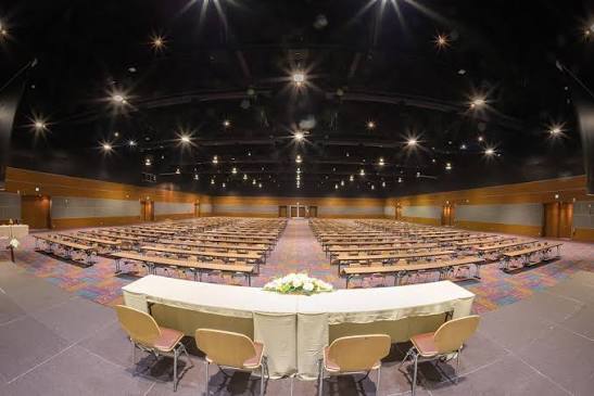 A vast dark exhibition hall set with long rows of banquet tables and gold-sashed chairs stretching toward a curtained stage, spotlights and a starlit ceiling rig overhead.