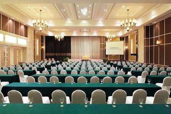 A ballroom set classroom-style with rows of covered tables and chairs facing a stage, a screen reading Imperial Hotel and Convention Centre beneath crystal chandeliers.