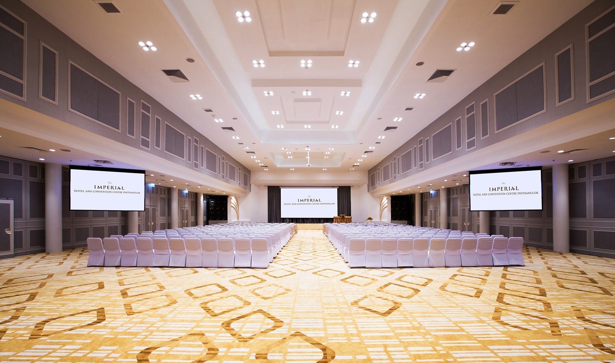 A ballroom set theatre-style with white chairs facing a stage and screens, the side and rear screens both reading The Imperial Hotel and Convention Centre Phitsanulok.