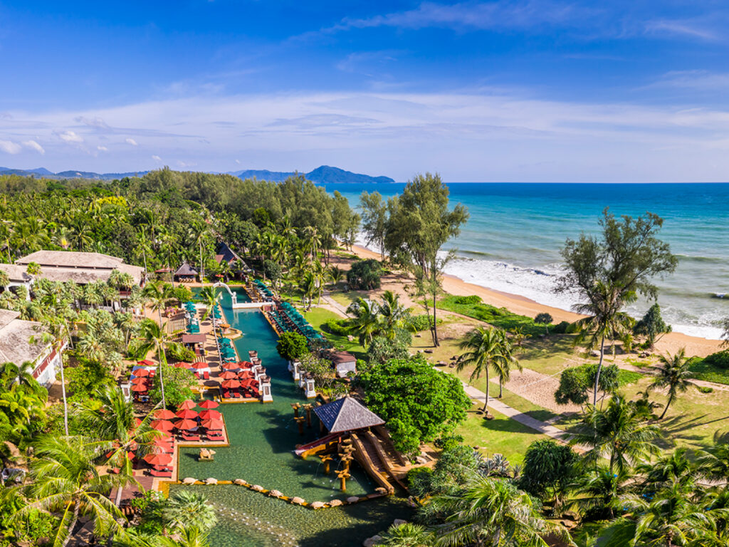 An aerial view along the resort's lagoon-style pool lined with red umbrellas and loungers, thatched-roof pavilions among palms, the beach and sea to the left.