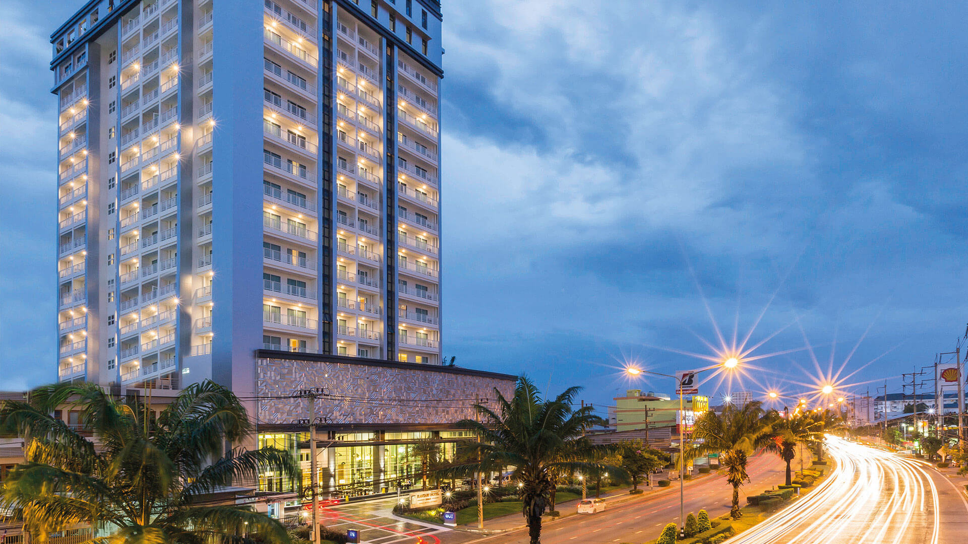 The hotel tower at dusk from across the main road, its facade lit and balconies illuminated, palms in the forecourt and light trails from passing traffic.