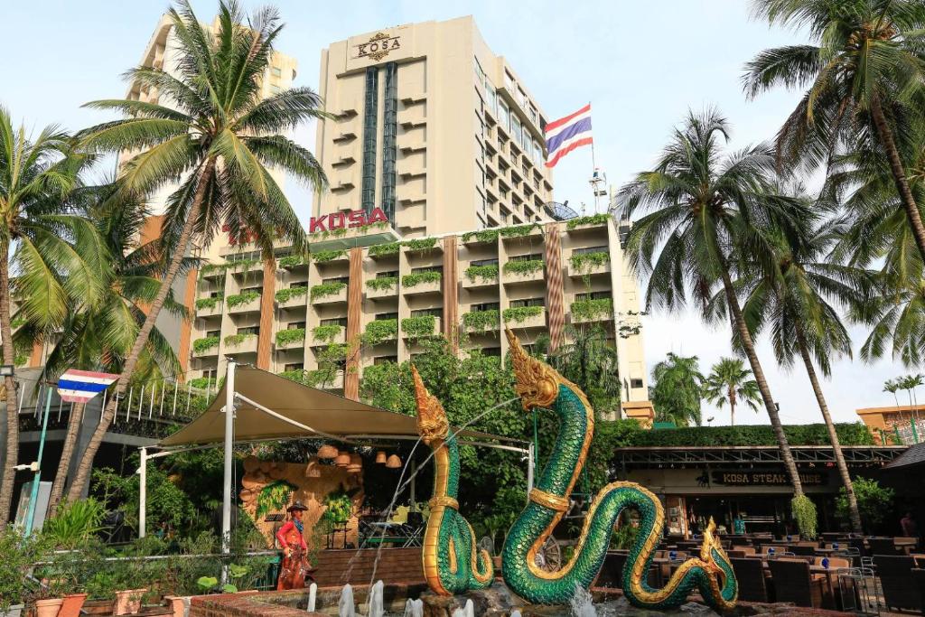 The hotel tower behind a Naga fountain sculpture and steak-house terrace, KOSA lettering on the tower and a Thai flag flying above, palm trees framing the entrance.