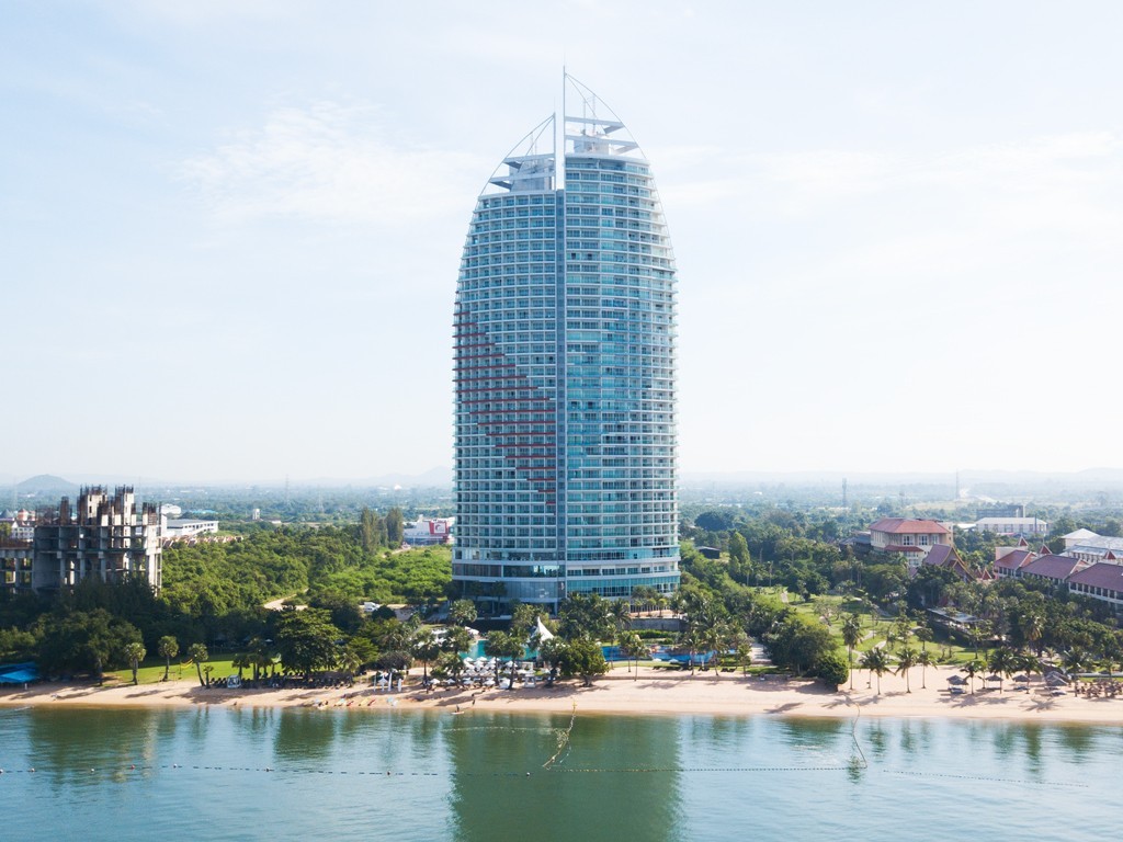 A tall sail-shaped glass tower on the beachfront, its curved facade rising above the shoreline with the sea in the foreground.