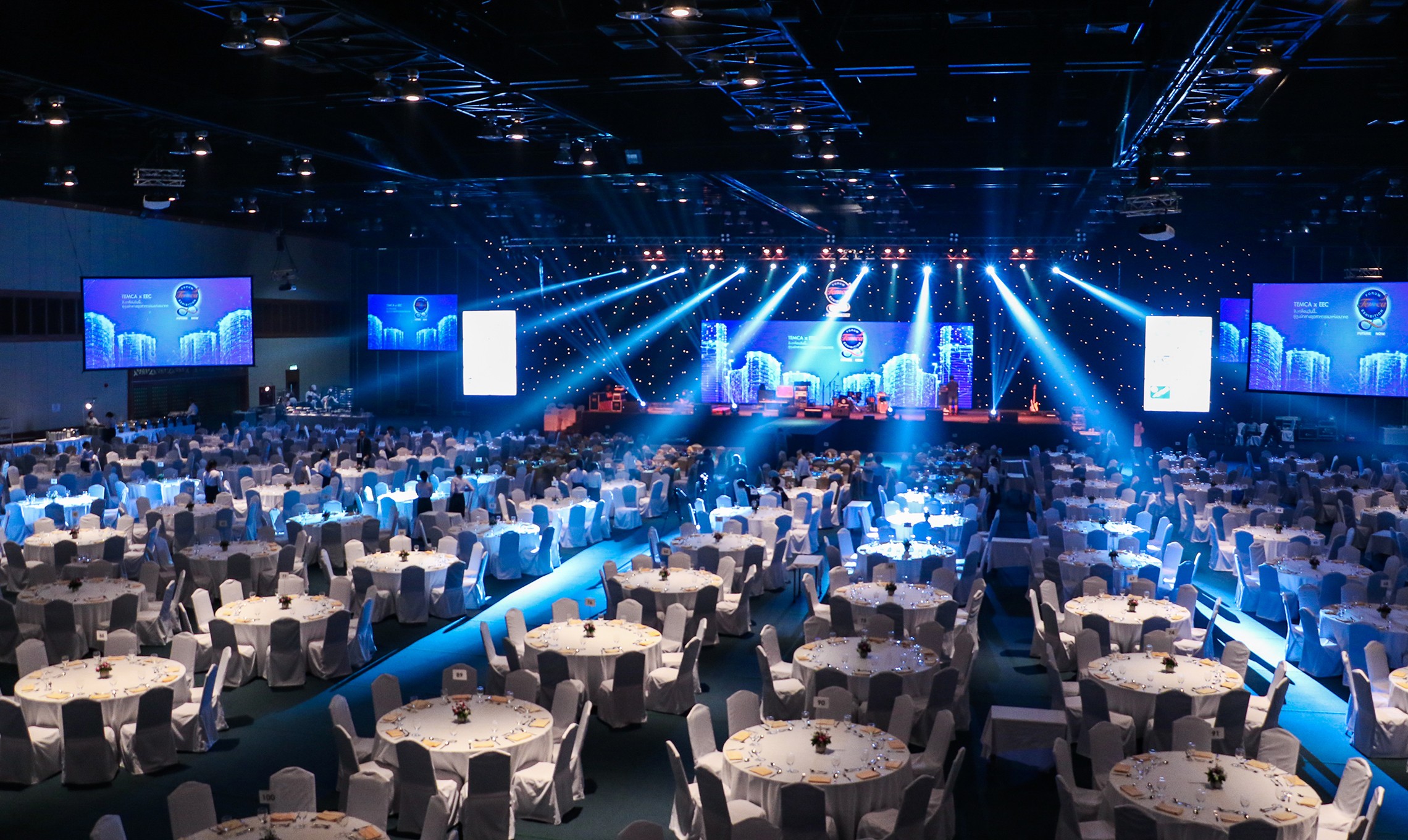 A banquet hall set with hundreds of round tables and white chair covers beneath stage lighting rigs, screens either side and a live band performing on the raised stage.