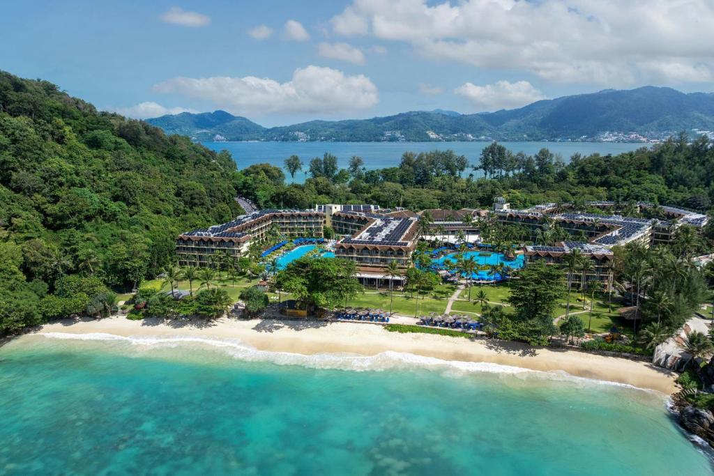An aerial view of the resort's solar-panelled rooftops and two pools set into a wooded hillside above a curving cove beach, with Patong's bay and hills beyond.