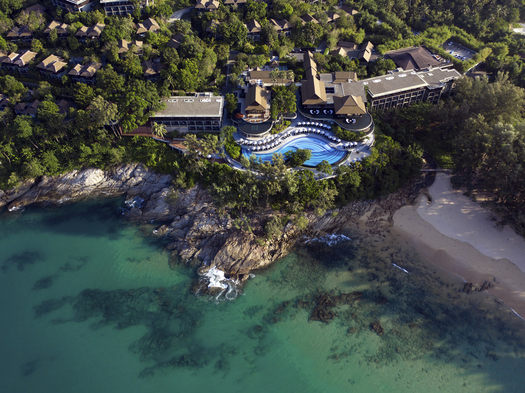 An aerial view of the resort's curved pool on a rocky headland, waves breaking on the rocks below and a sandy cove beach to the right.
