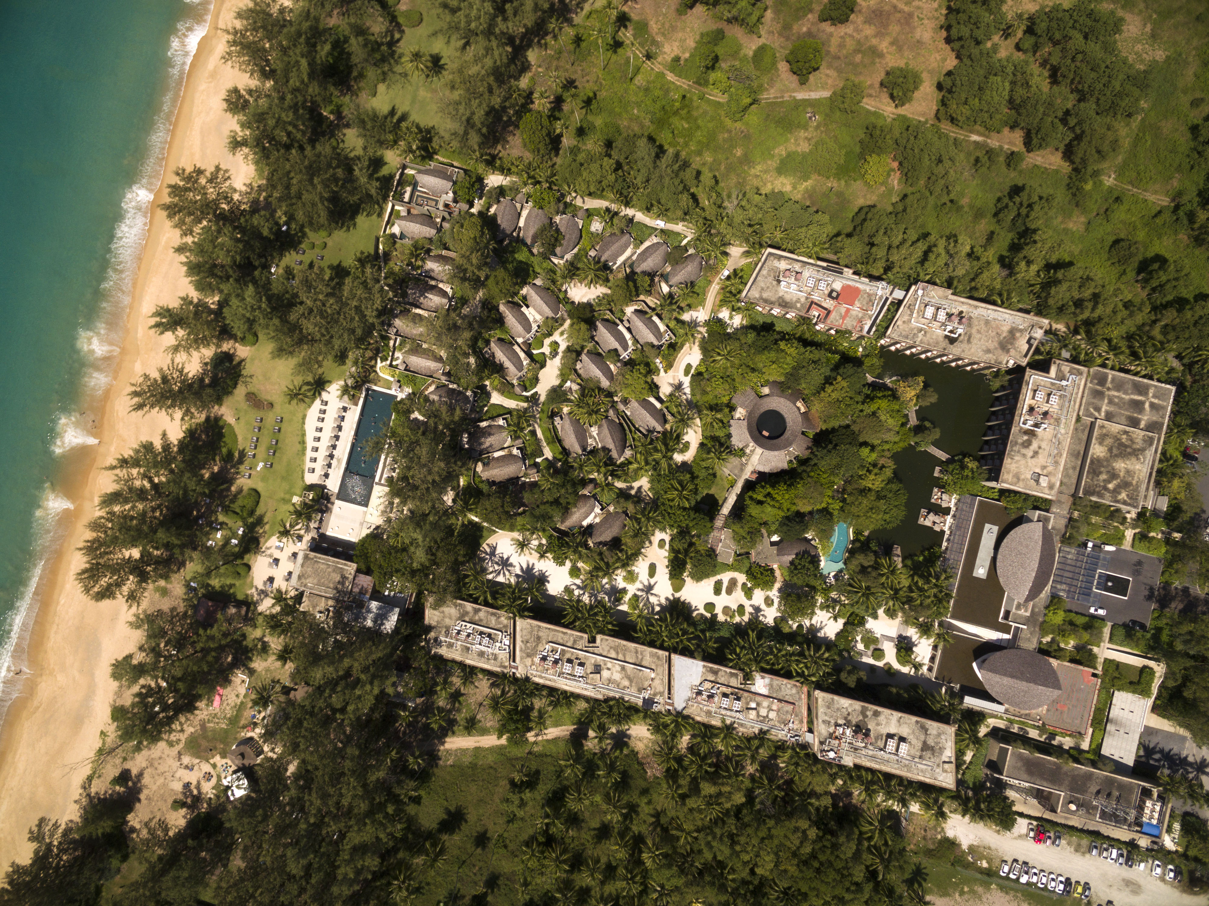 A straight-down aerial view of the resort's circular thatched villas clustered around a dark lily pond, with the main building and a further pool to the right.