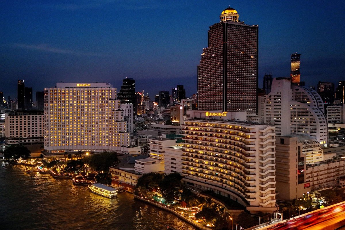 The hotel's two riverside wings lit at night on the Chao Phraya, its name in Thai and English along the rooflines, a boat at the pier below.