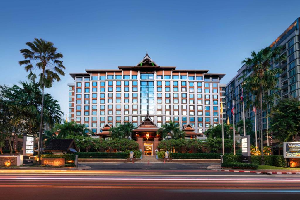 The hotel at dusk behind its forecourt, a tiered Lanna roof crowning the block, palms either side and light trails on the road in front.
