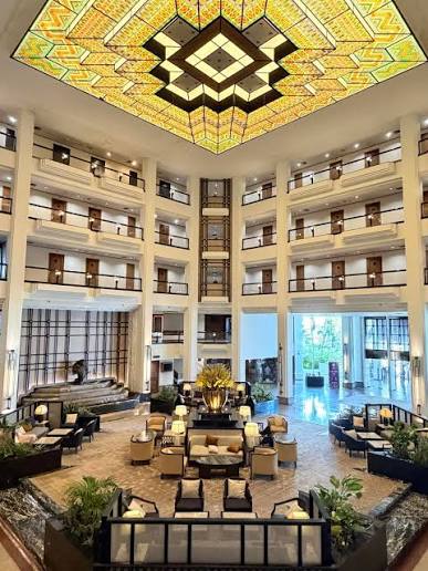 A four-storey atrium lobby lounge with a large geometric stained-glass skylight overhead, balconied rooms rising on all sides above a marble-floored seating area.