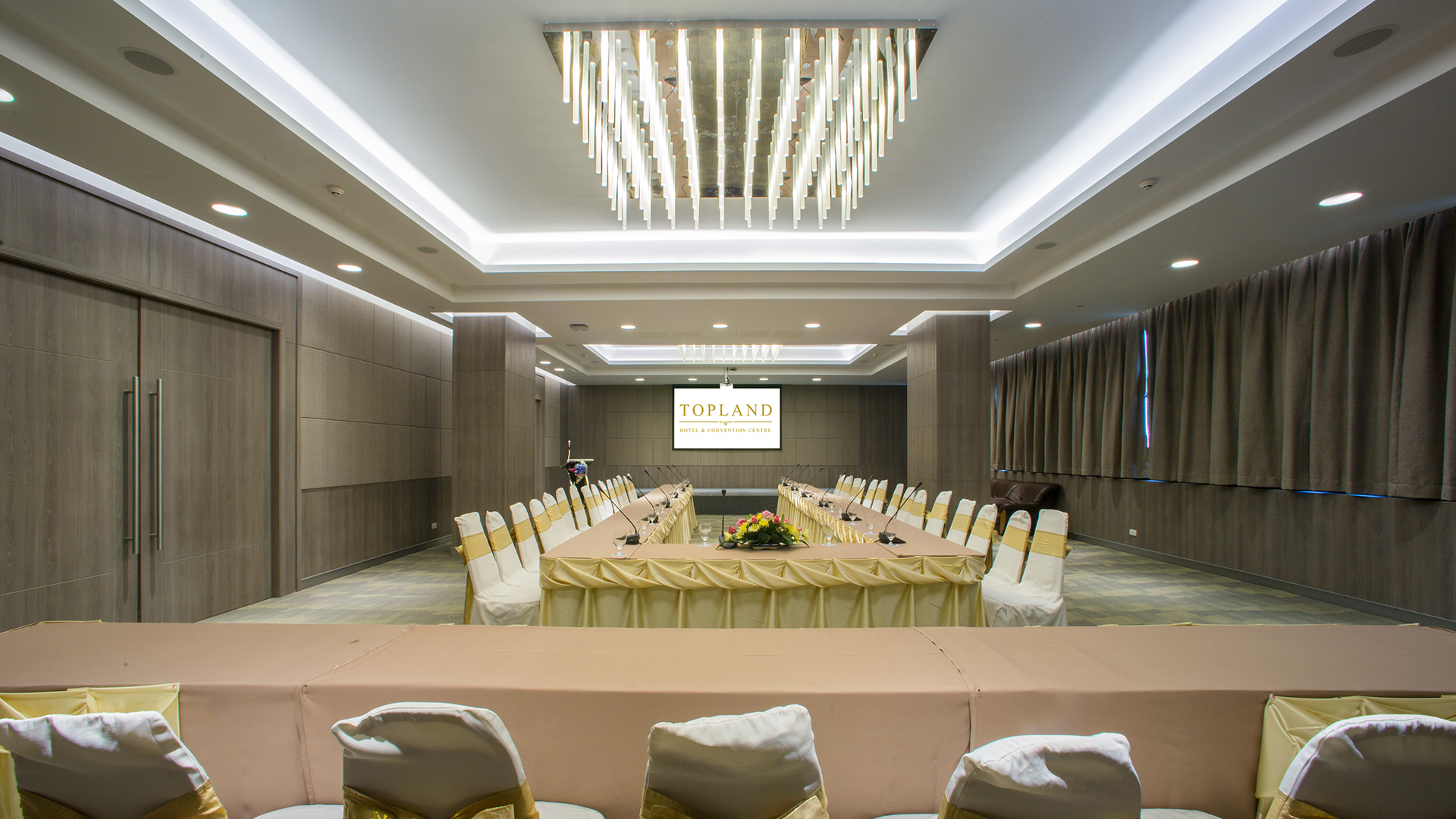 A meeting room set boardroom-style with a long U-shaped table and gold-sashed chairs, a screen at the front reading Topland Hotel & Convention Centre beneath a sculptural ceiling light.