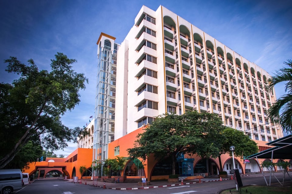 The hotel's orange and cream tower on a street corner in Surat Thani town, a glazed lift shaft rising beside arched ground-floor arcades and mature trees along the road.