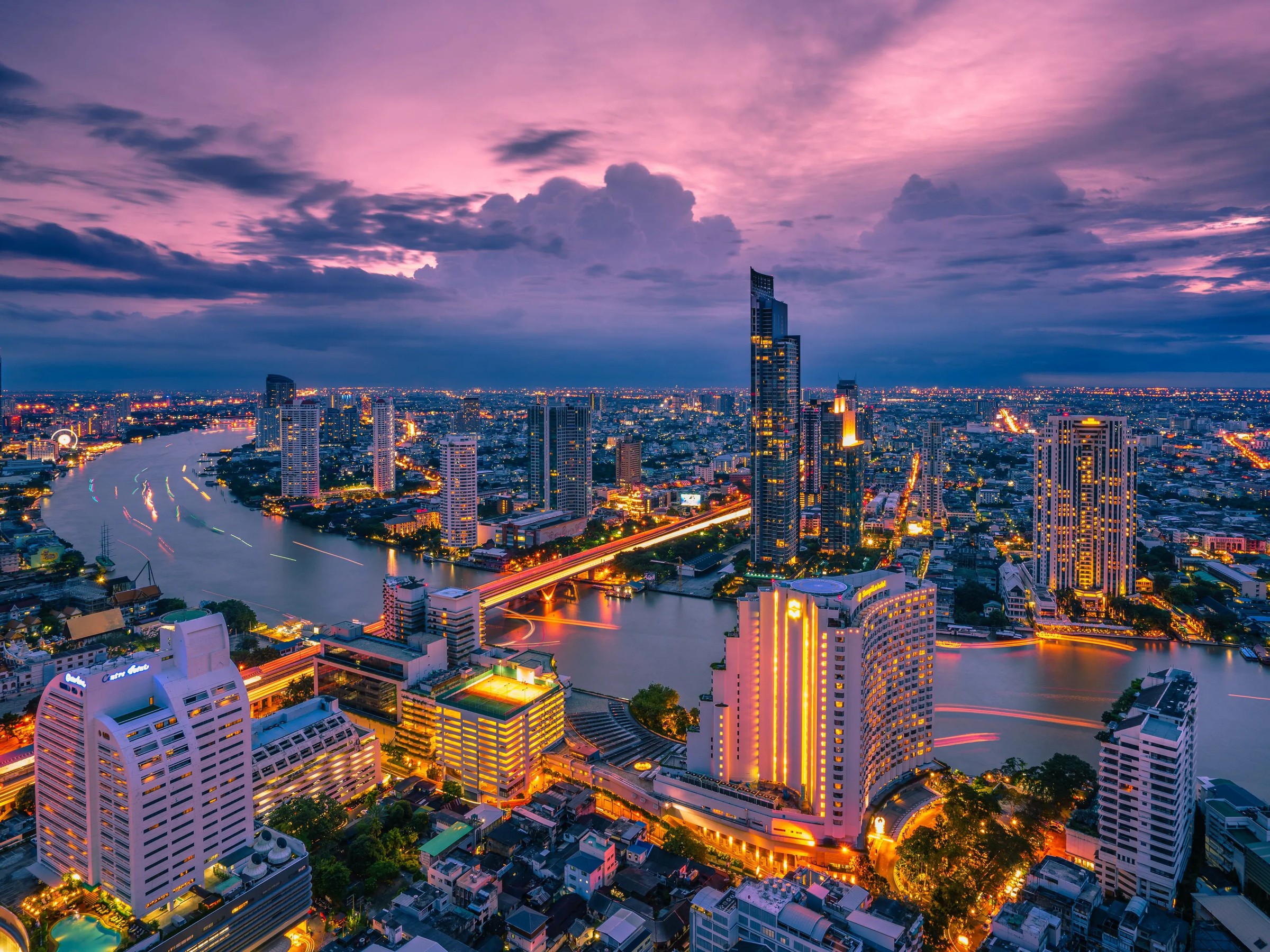 The Chao Phraya curving through Bangkok at dusk, the city lit beneath a violet sky