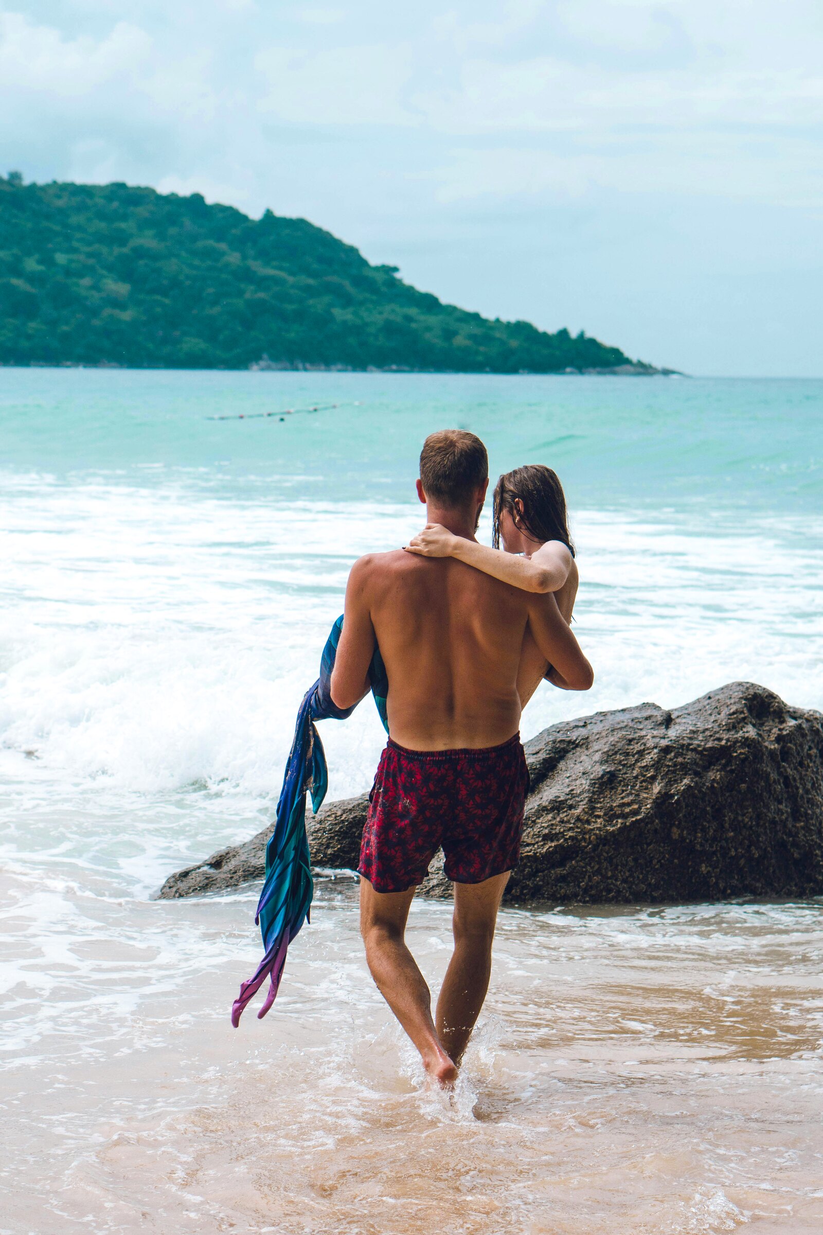 A man carrying a woman through the shallows on a beach, a forested headland behind