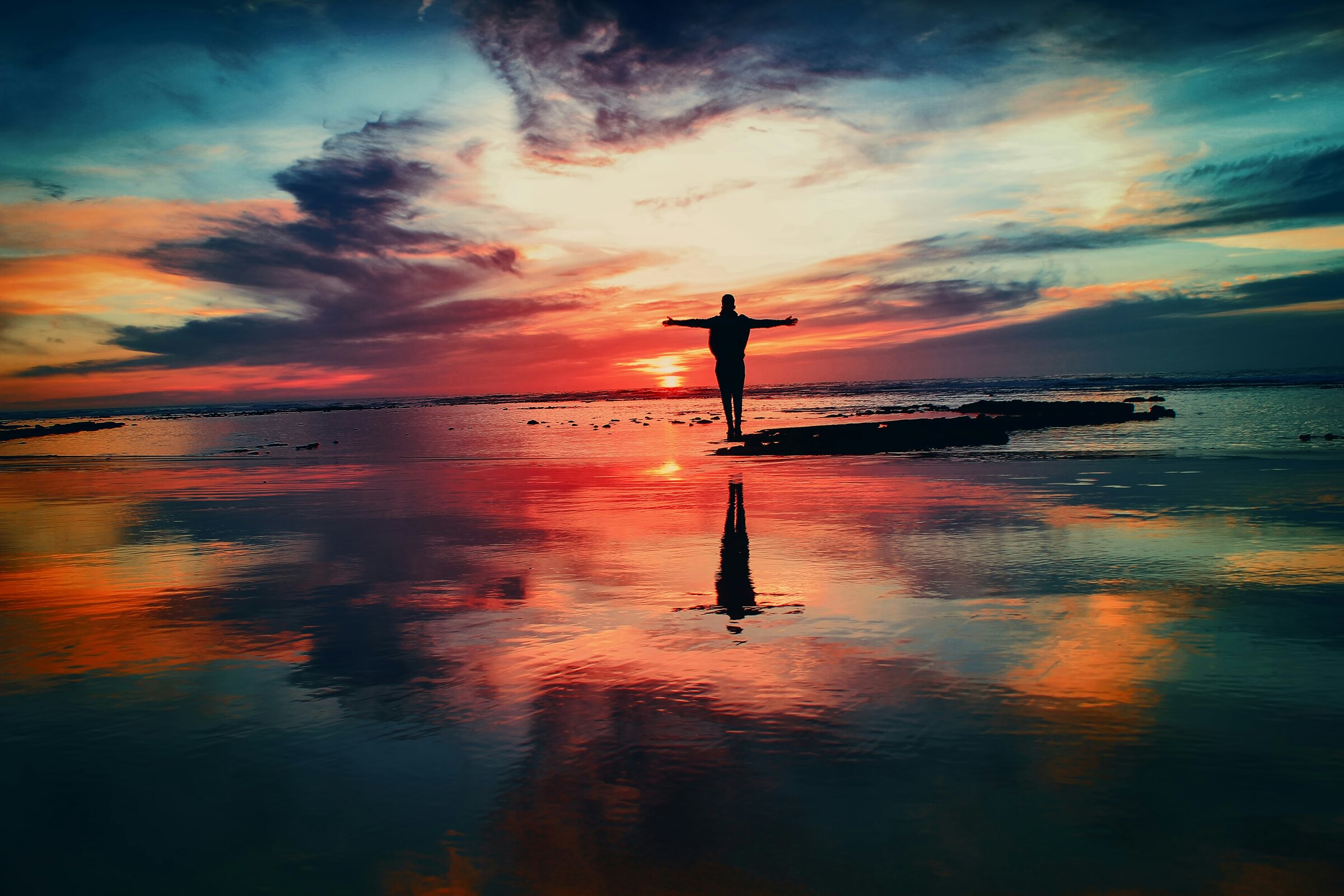 A figure with arms outstretched on a wet beach at sunset, reflected in the shallows