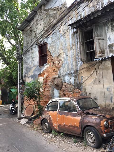 A weathered Talat Noi shophouse, its plaster peeling back to brick, with a rusting vintage car parked at the foot of the wall