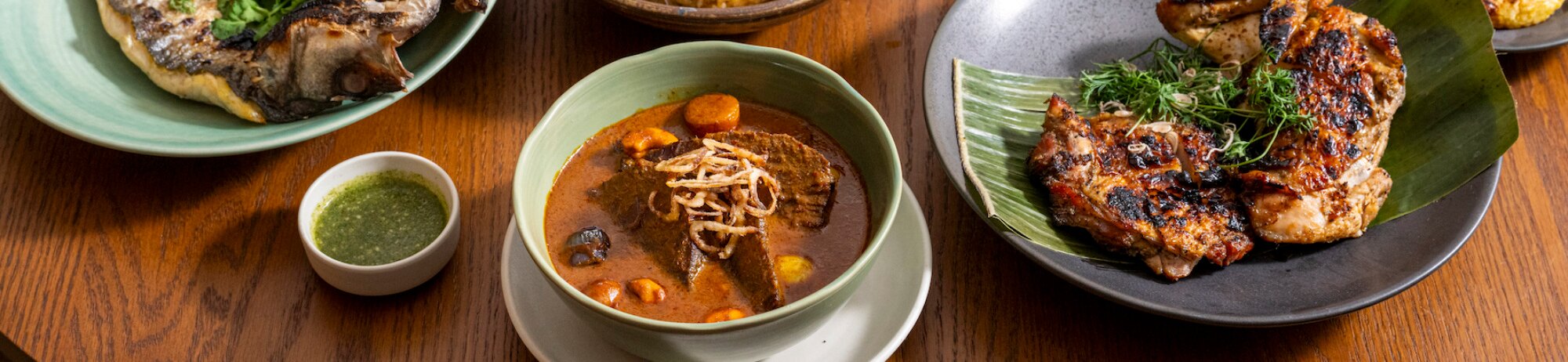 A spread of Thai dishes on a wooden table — fried fish, a bowl of massaman with fried shallots, grilled chicken on a banana leaf