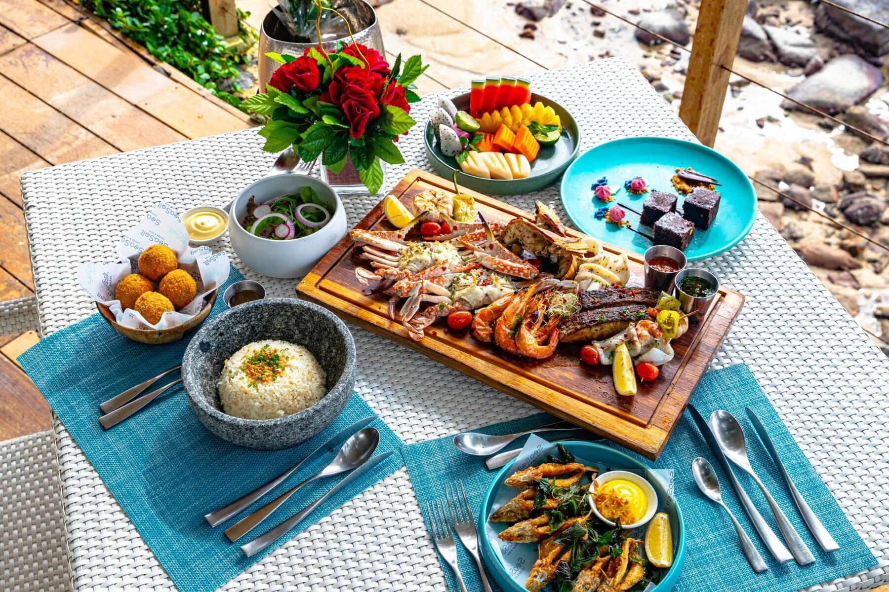 A seafood board on a beachside table — crab, prawns, squid and grilled fish, rice, fruit and a plate of dessert